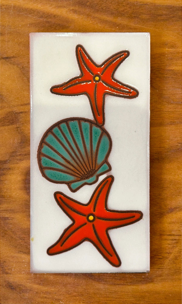 Decorative tile with red starfish and teal shell on a white background, placed on a wooden surface.