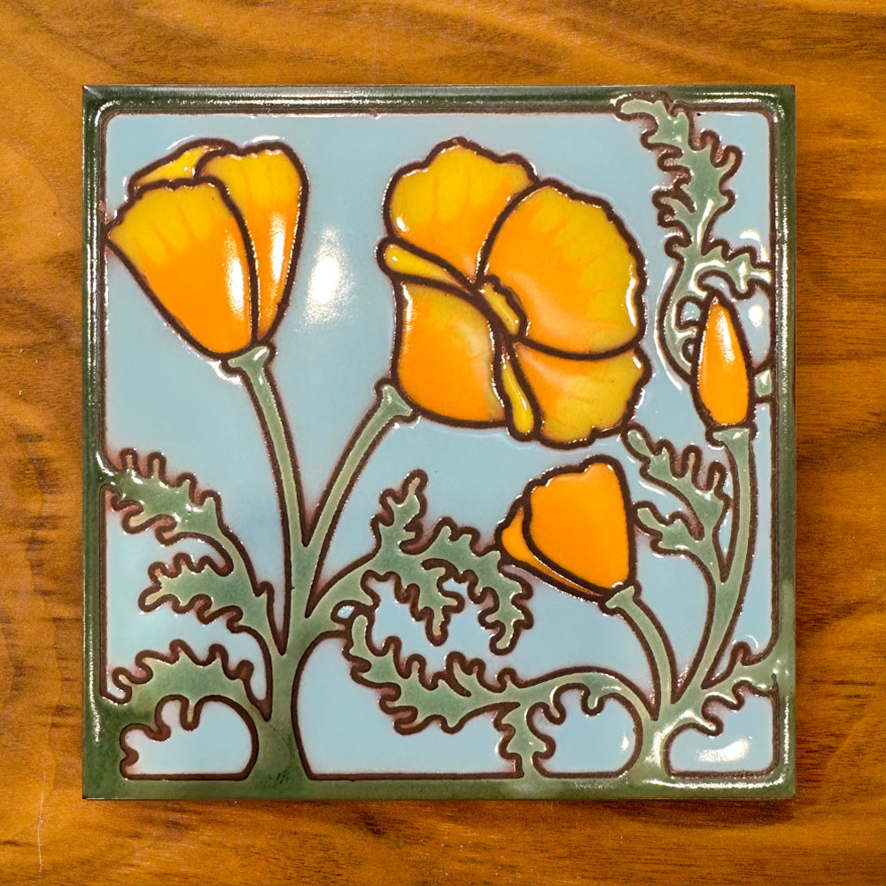 A ceramic trivet with a California poppies tile design in orange and green colors on a blue background, cork backed, with a hole at the top for hanging.