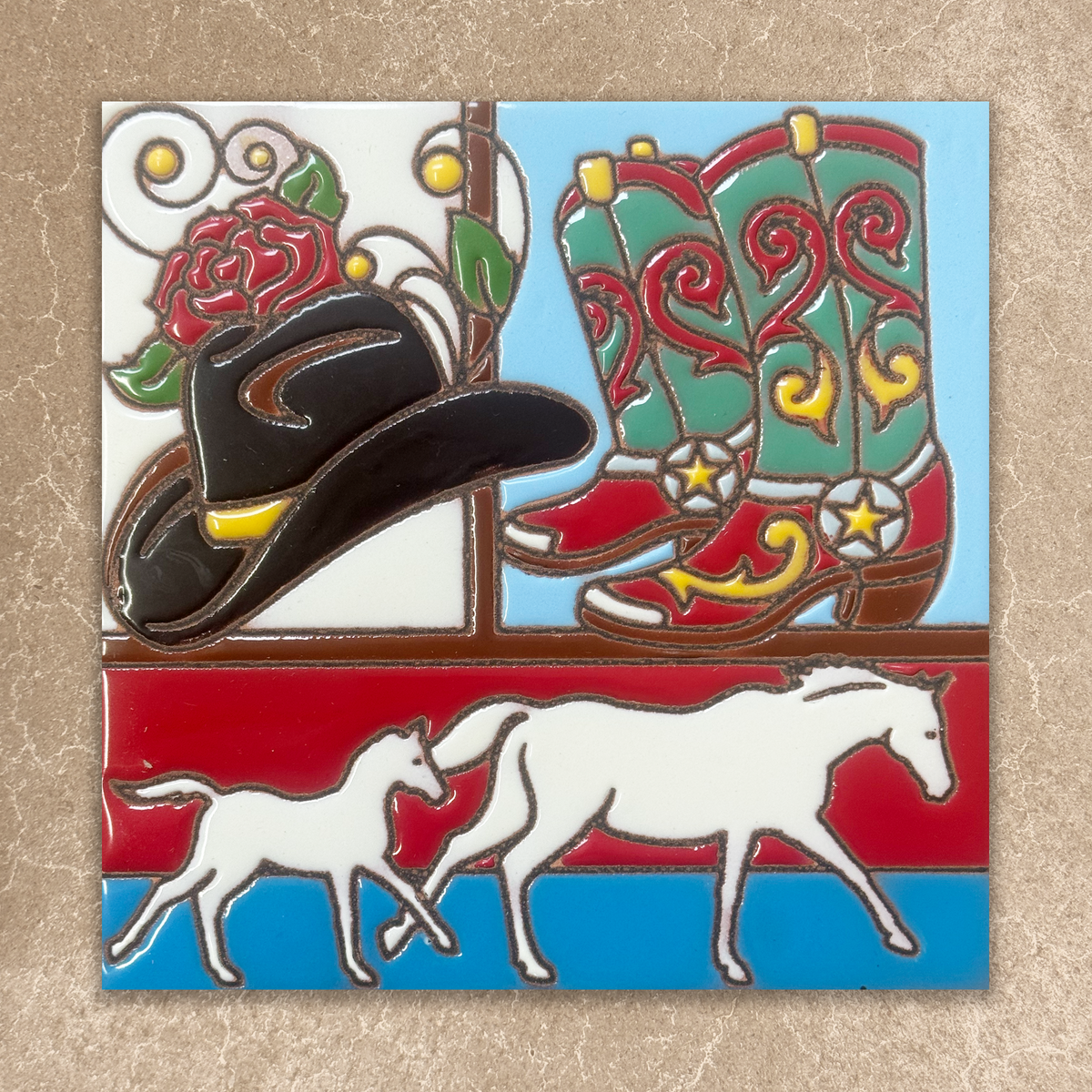 A square ceramic tile featuring a western-themed design with a black cowboy hat, red and green cowboy boots, a red rose, and white horses on a red and blue background, displayed on a textured beige surface.