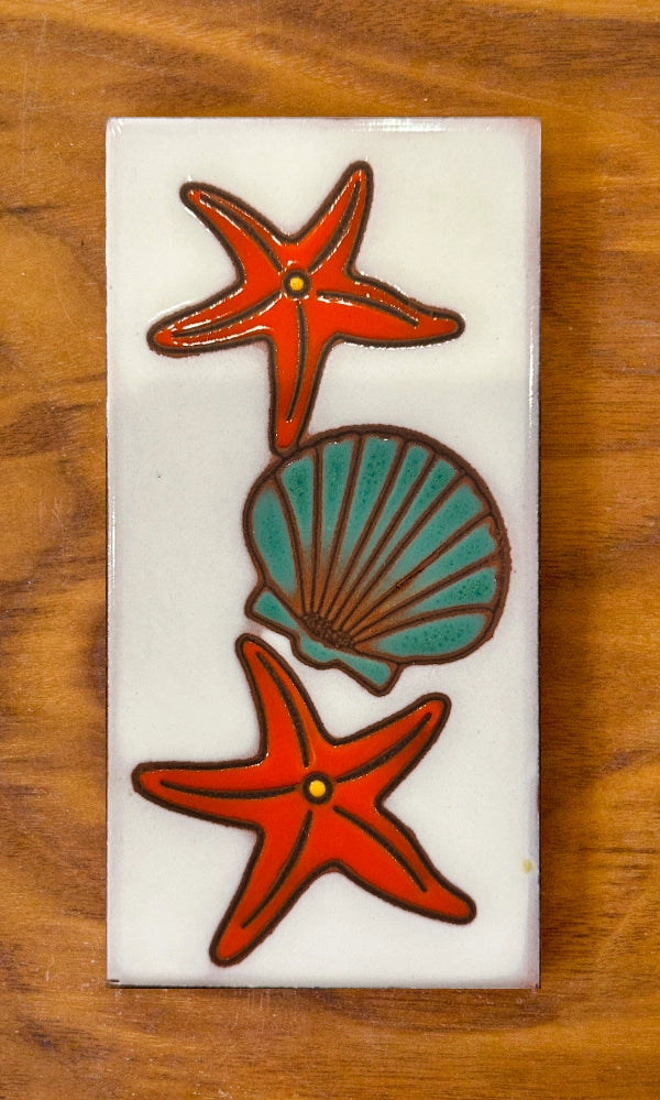Decorative tile with two red starfish and a blue shell on a white background, placed on a wooden surface.