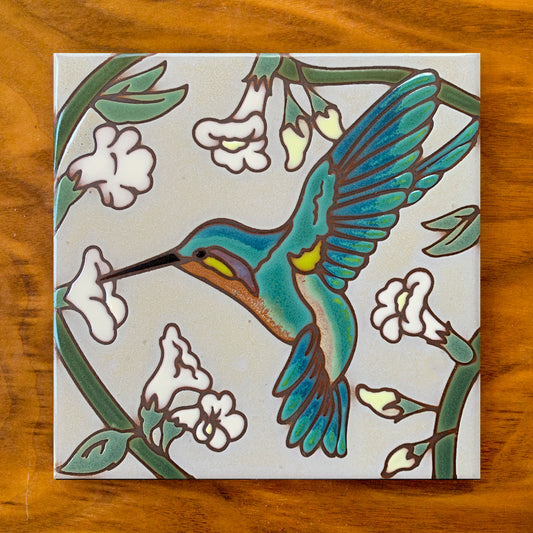 A hand-glazed ceramic tile art trivet featuring a hummingbird with white flowers design on a wooden surface.