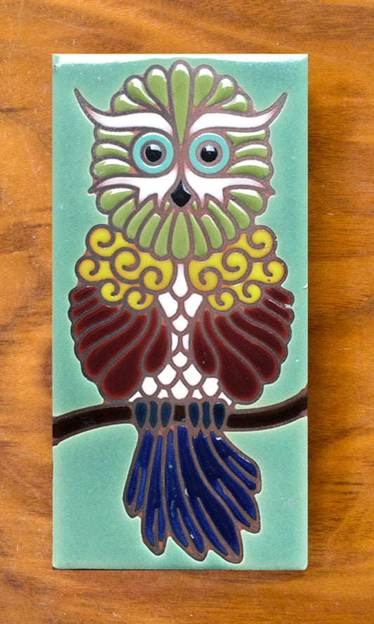 A hand glazed ceramic tile shaped like an owl, with a green, blue, and brown color scheme, and a cork backed for coaster use.