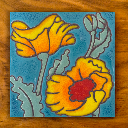 A square ceramic tile featuring a stylized poppy design with yellow and orange petals, a red center, and textured light blue-green leaves on a turquoise blue background, displayed on a wooden surface.