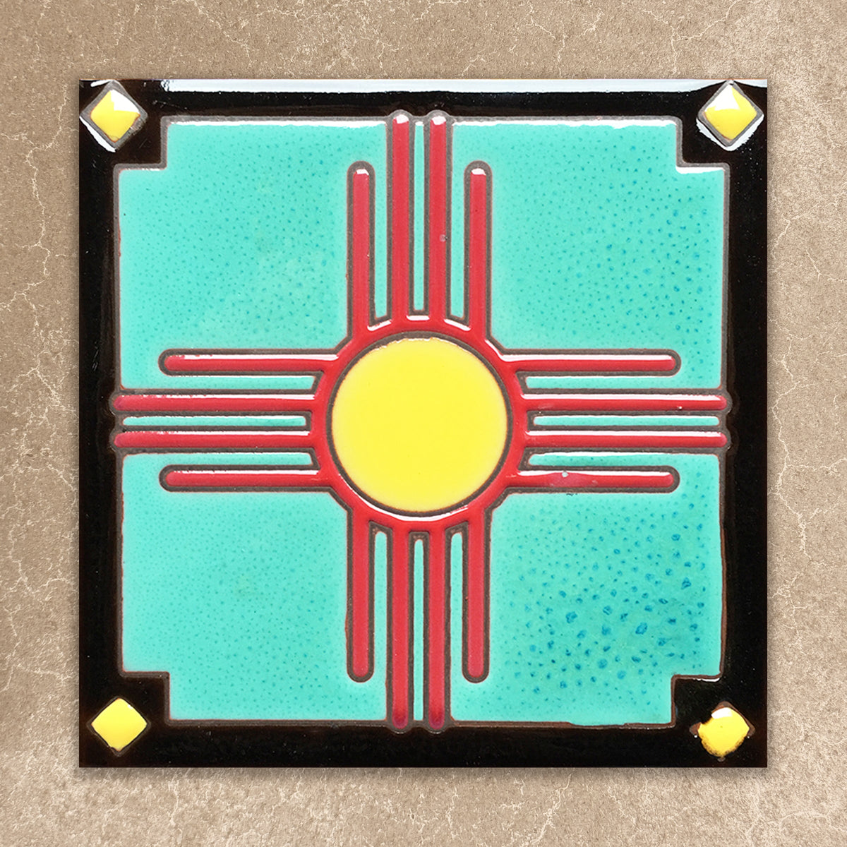 Decorative tile with a red and yellow zia symbol on a turquoise background, framed by black border with gold accents.