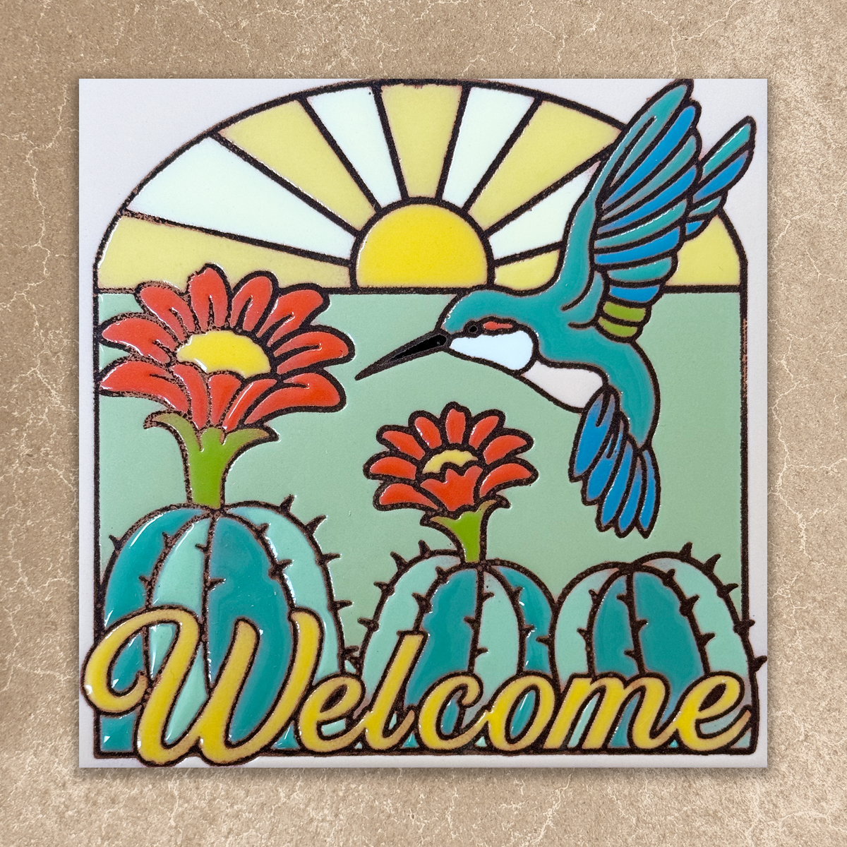 A square decorative tile or plaque featuring a colorful desert-style illustration with a hummingbird, cacti, flowers, a sun, and the word “Welcome” in large yellow script. The tile is centered on a textured beige background.