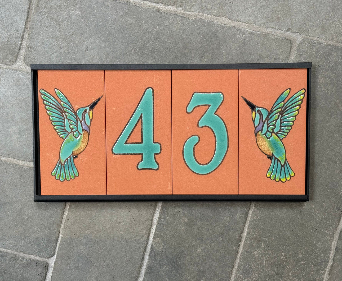 Decorative house number sign with hummingbirds on a stone pavement background