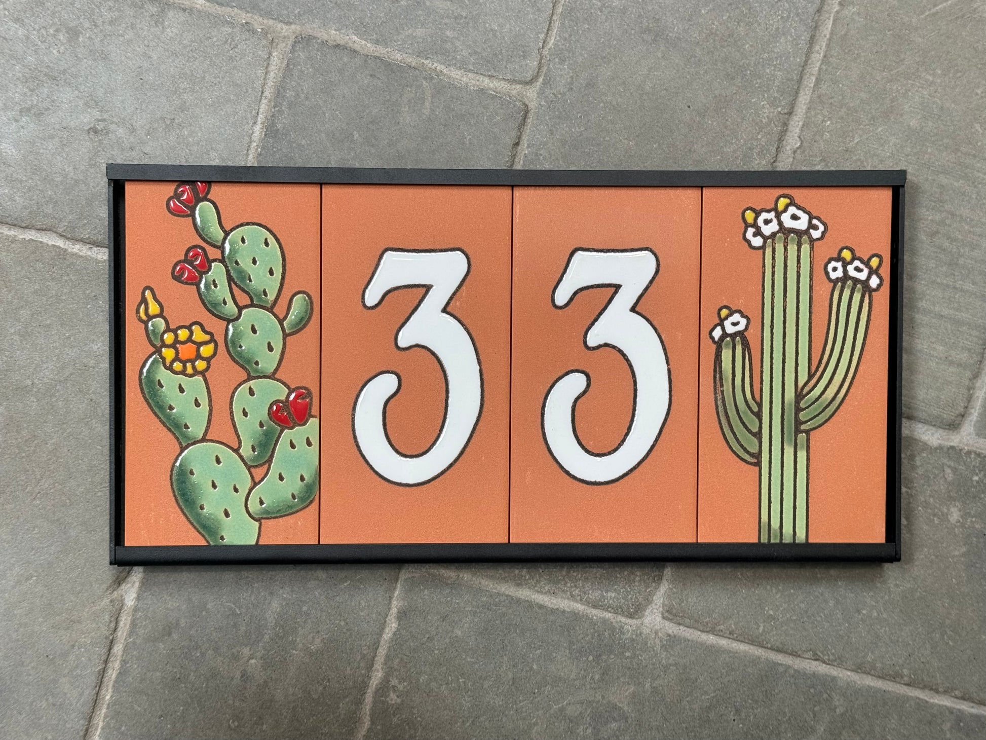 Decorative house number sign with cacti on an orange background, mounted on a stone wall.