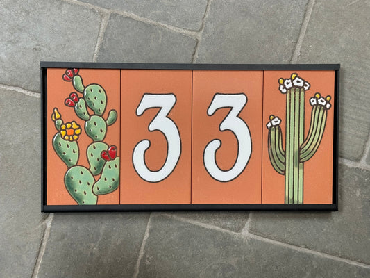 Decorative house number sign with cacti on an orange background, mounted on a stone wall.