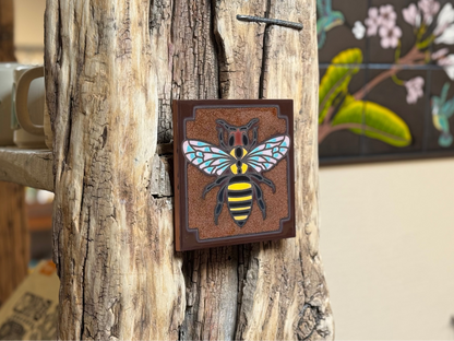 Decorative bee painting on a wooden surface with mugs and a floral painting in the background.