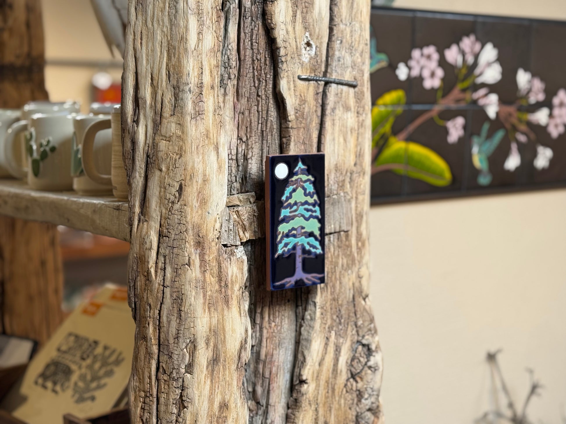Decorative tree magnet attached to a wooden surface with a blurred background of mugs and artwork.