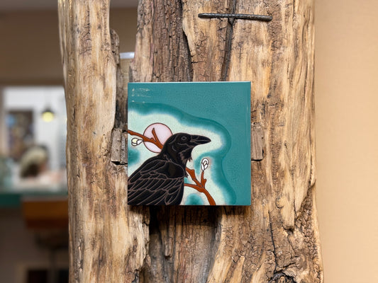 Small square painting of a black bird on a branch attached to a wooden surface.