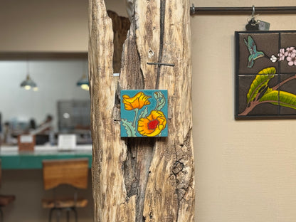 Decorative tile with floral design attached to a wooden surface, with a blurred indoor setting in the background.