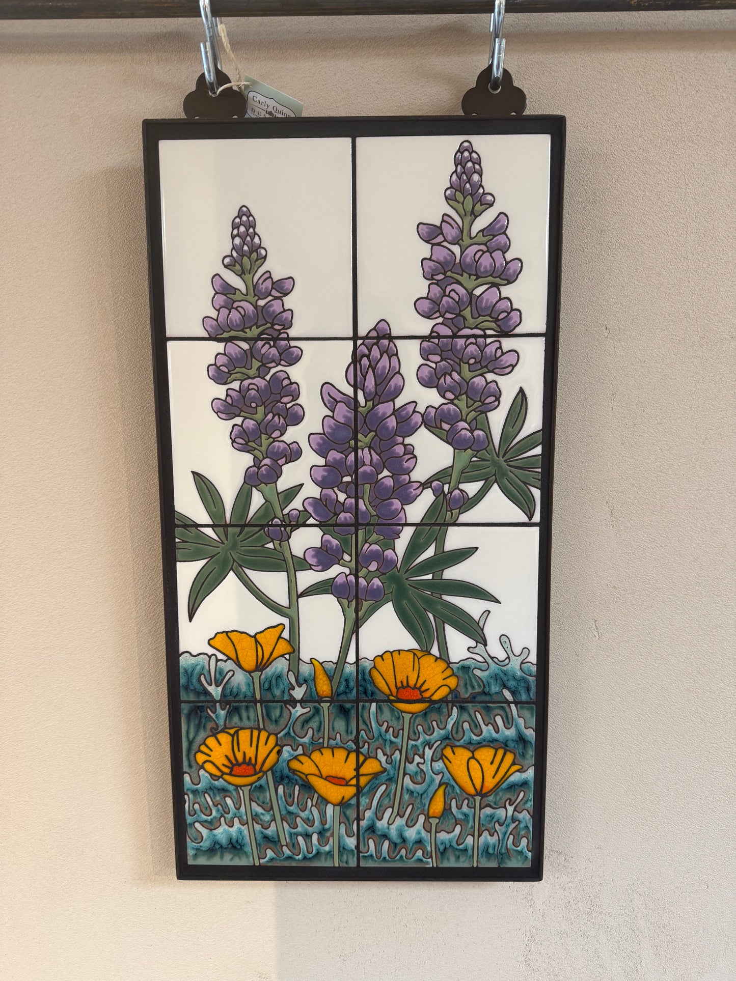 A rectangular ceramic tile wall hanging with a black frame, featuring purple lupines and yellow poppies on a white background with blue-green foliage and ground, displayed against a beige wall.