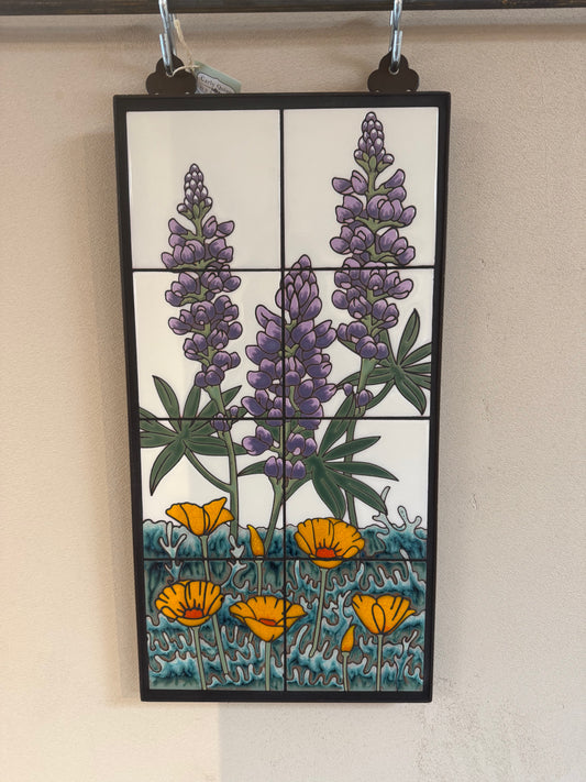 A rectangular ceramic tile wall hanging with a black frame, featuring purple lupines and yellow poppies on a white background with blue-green foliage and ground, displayed against a beige wall.