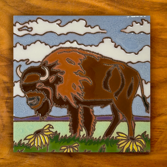 A square ceramic tile featuring an artistic depiction of a brown bison in a grassy field with yellow flowers, under a blue sky with white clouds, displayed on a wooden surface.
