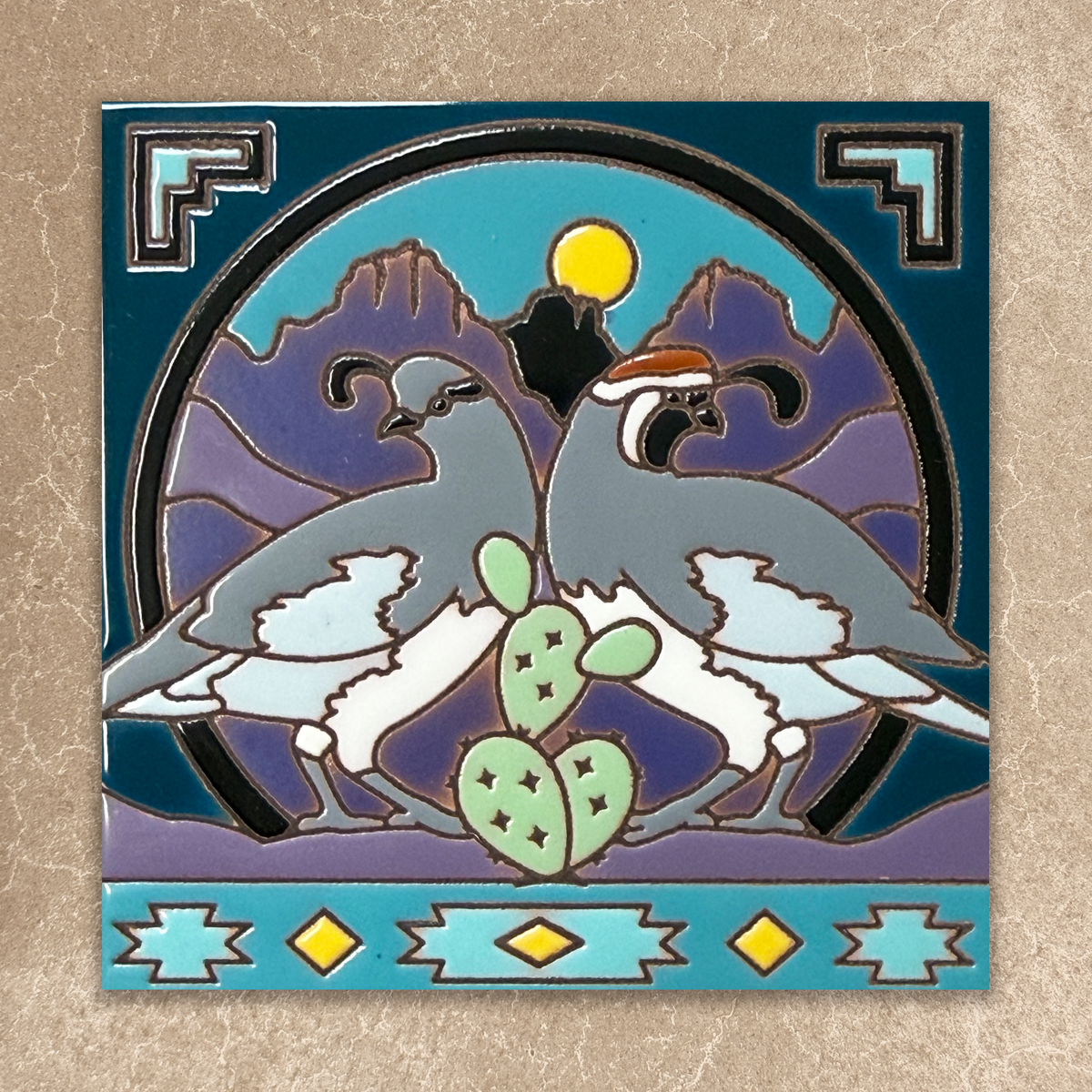 A square enamel pin featuring two gray and white quail birds with a green prickly pear cactus, set against a purple mountain background with a yellow sun and black circular frame. The pin is mounted on a dark teal border with geometric patterns, shown on a textured beige background.