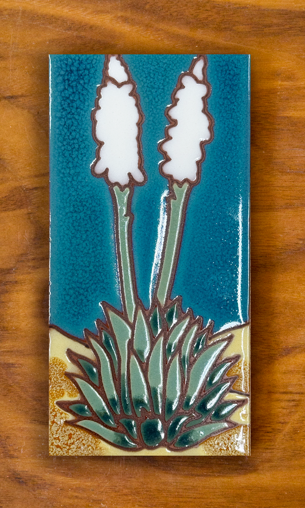 A rectangular ceramic tile featuring a stylized desert plant design with two white flower spikes on green stems and a textured gold base, set against a teal blue background, displayed on a wooden surface.