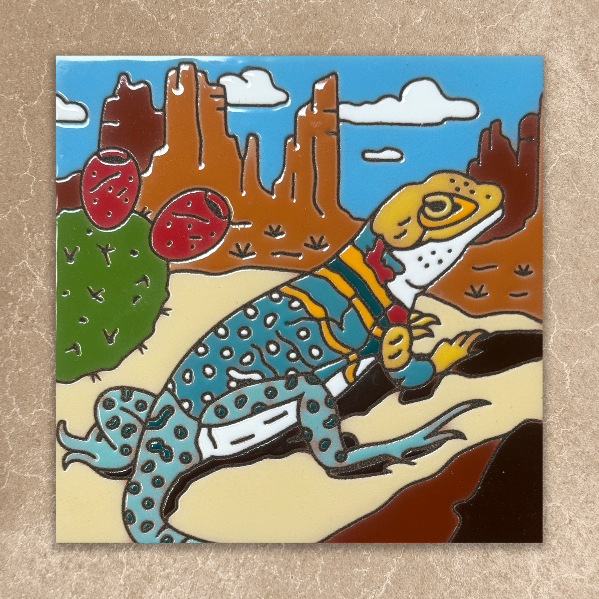 Colorful ceramic tile of a lizard in a desert landscape with cacti and mountains.